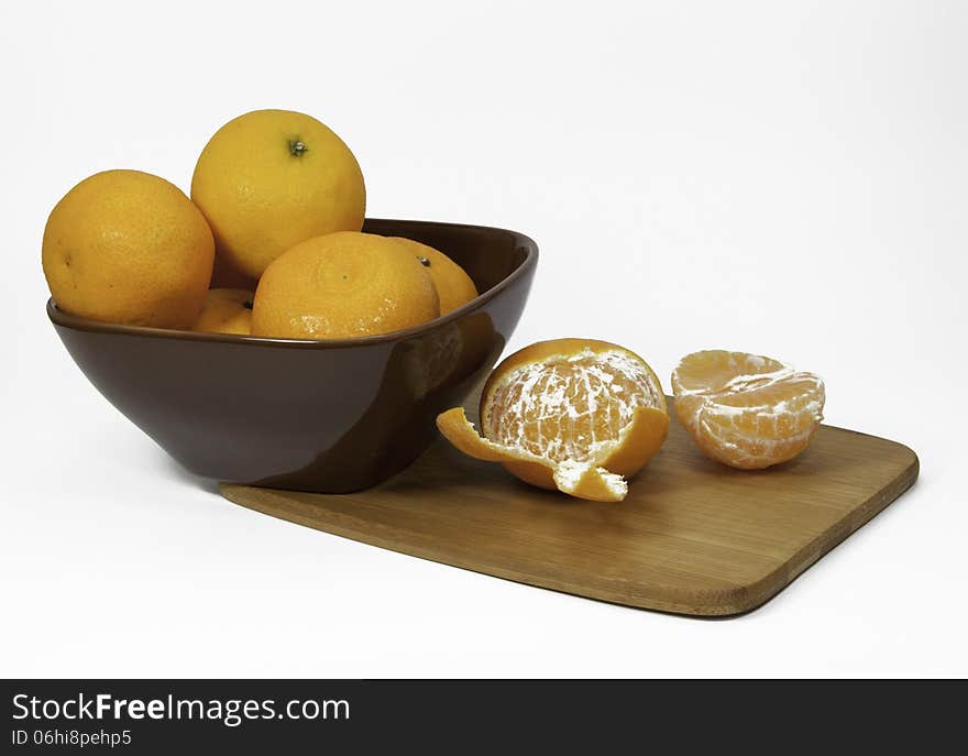 Clementine oranges in a brown bowl and unpeeled on a bamboo cutting board against a white background. Clementine oranges in a brown bowl and unpeeled on a bamboo cutting board against a white background