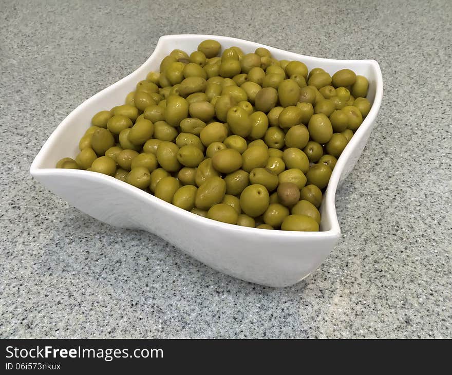 Aerial view of a green olives contained in a white square bowl resting on a kitchen counter top. Aerial view of a green olives contained in a white square bowl resting on a kitchen counter top