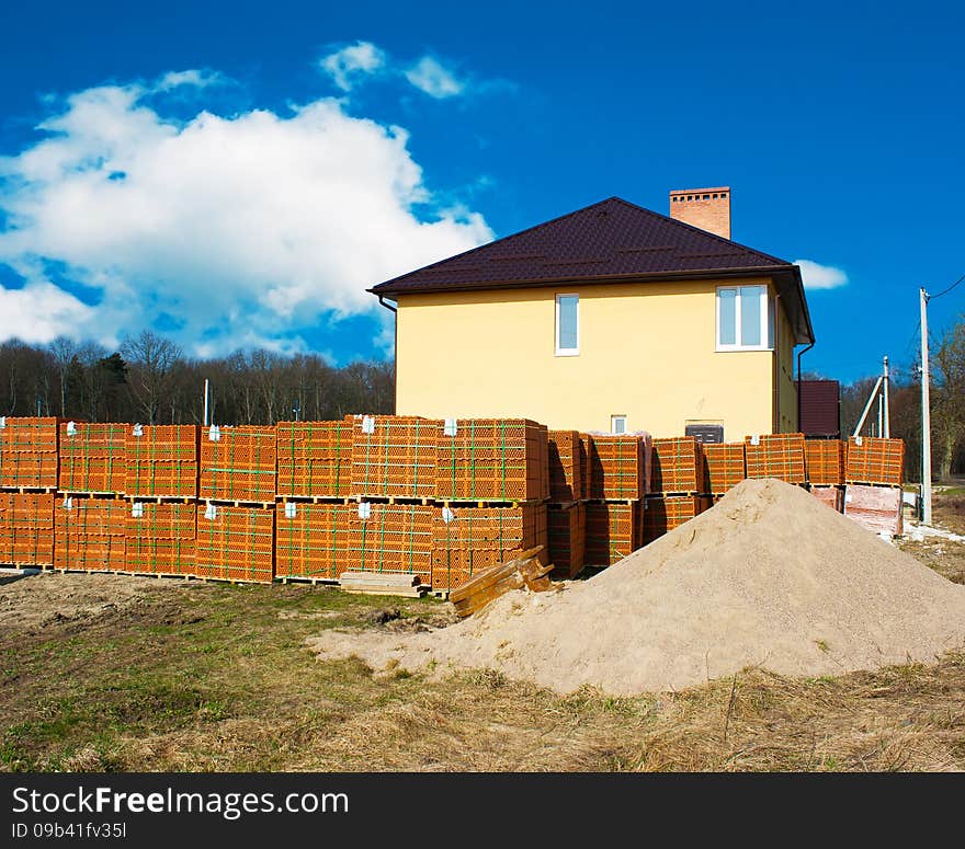 Stacks of red bricks for the construction and new yellow house with blue cloudy sky. Stacks of red bricks for the construction and new yellow house with blue cloudy sky