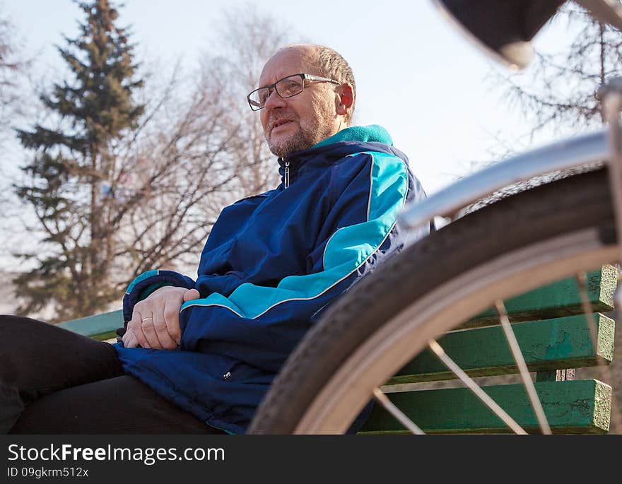 Portrait of eldely man wearing glasses, black trousers and a blue jacket sitting on a bench near his bicycle in a city park. Portrait of eldely man wearing glasses, black trousers and a blue jacket sitting on a bench near his bicycle in a city park