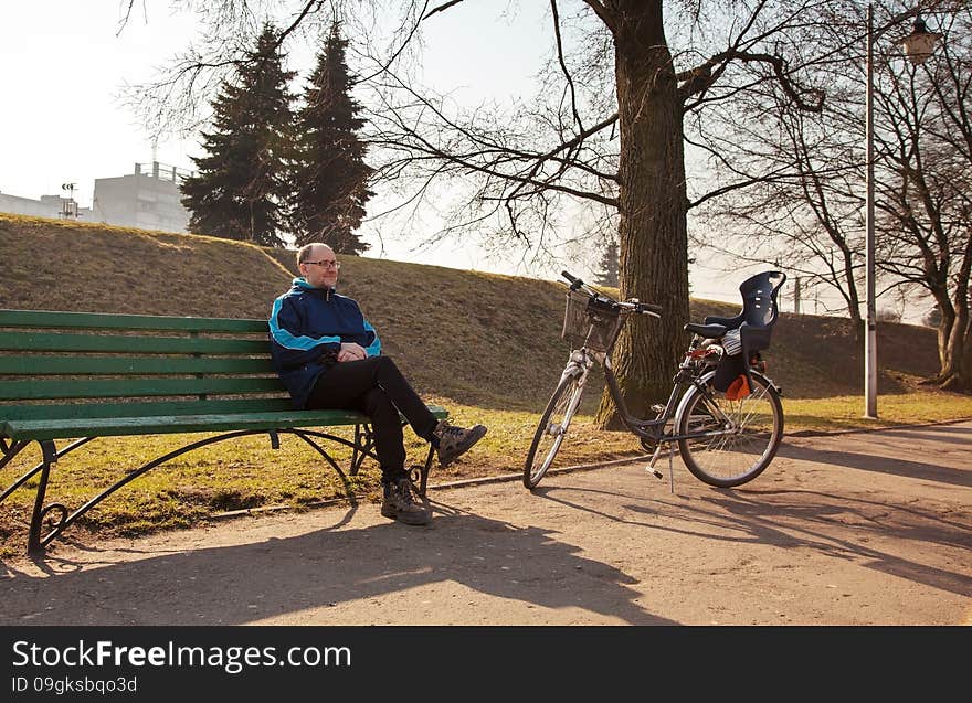 Elderly man wearing glasses, black trousers and a blue jacket sitting on a bench near his bicycle in a city park. Elderly man wearing glasses, black trousers and a blue jacket sitting on a bench near his bicycle in a city park