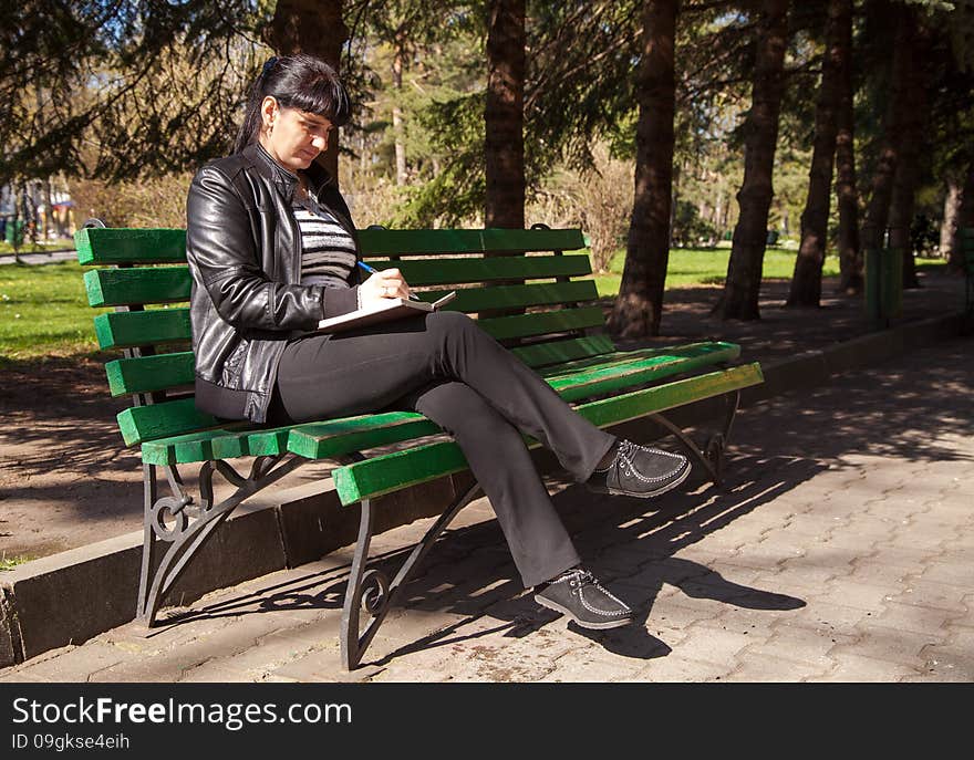 Young beautiful woman in a black leather jacket and black pants sitting on a park bench writing a pen in notebook on sunny spring day. Young beautiful woman in a black leather jacket and black pants sitting on a park bench writing a pen in notebook on sunny spring day