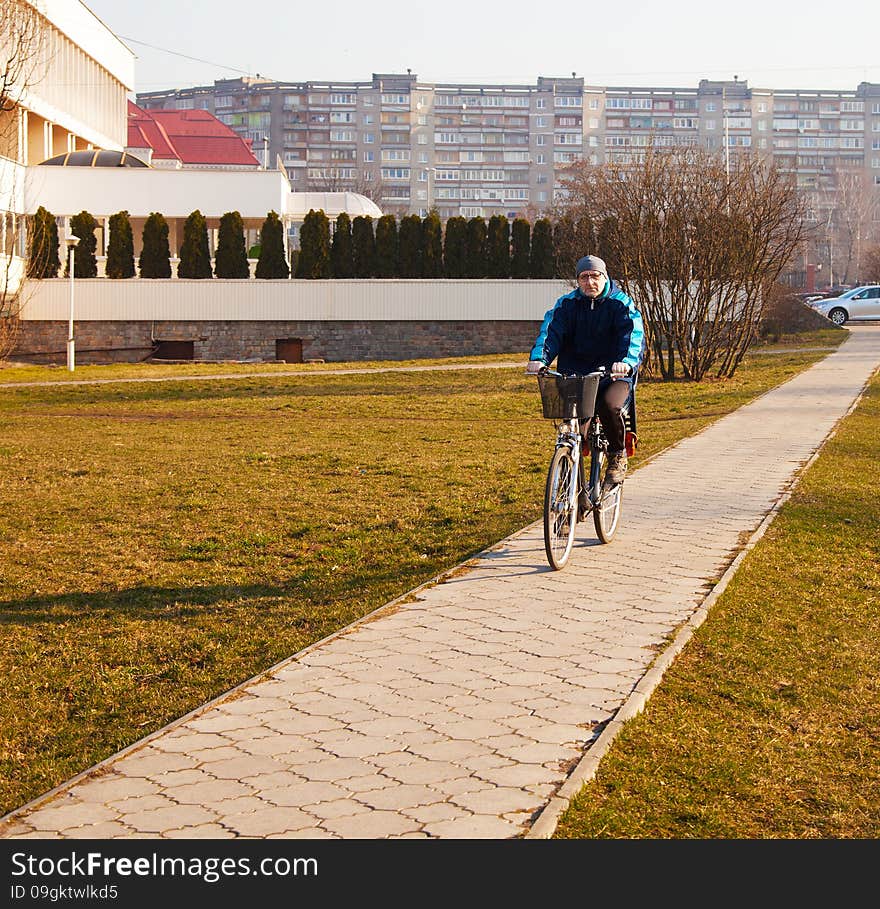 Elderly man wearing glasses, a blue jacket, black trousers and a gray hat riding a bicycle on the sidewalk of concrete tiles. Elderly man wearing glasses, a blue jacket, black trousers and a gray hat riding a bicycle on the sidewalk of concrete tiles