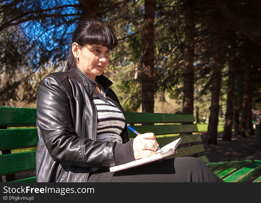 Young beautiful woman in a black leather jacket and black pants sitting on a park bench writing a pen in notebook. Young beautiful woman in a black leather jacket and black pants sitting on a park bench writing a pen in notebook