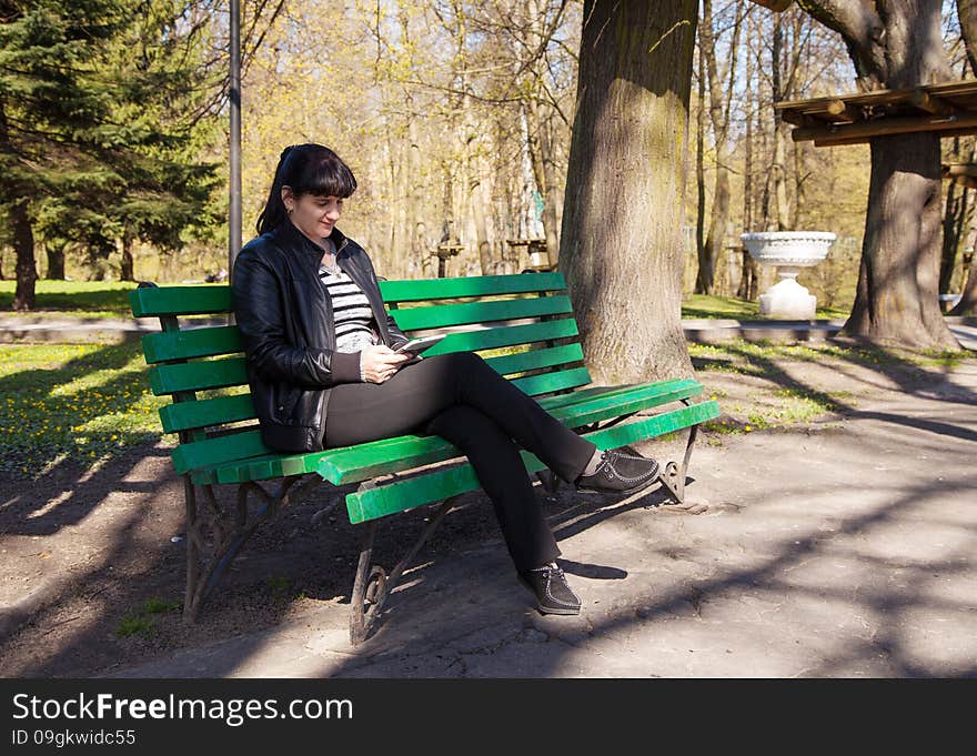 Young beautiful woman in a black leather jacket and black pants sitting on a park bench holding a tablet. Young beautiful woman in a black leather jacket and black pants sitting on a park bench holding a tablet