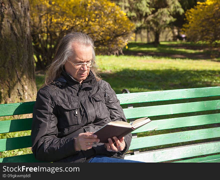 Haired elderly man in a black jacket and glasses reading a book sitting on a bench in city park on sunny spring day. Haired elderly man in a black jacket and glasses reading a book sitting on a bench in city park on sunny spring day