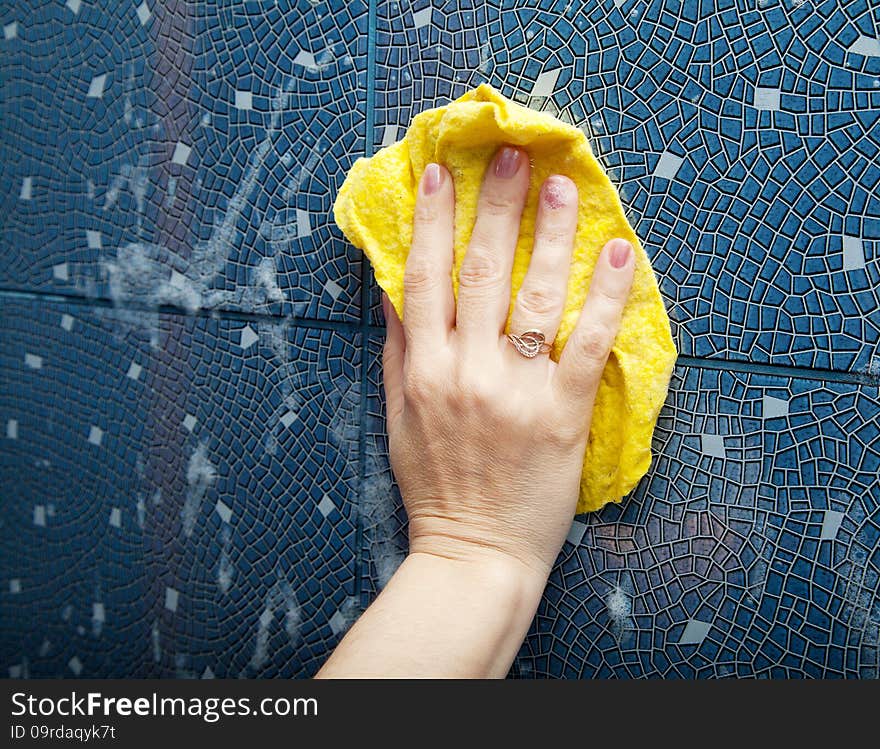 Female hand washes the blue tile on the wall with a yellow cloth lather in the bathroom closeup. Female hand washes the blue tile on the wall with a yellow cloth lather in the bathroom closeup