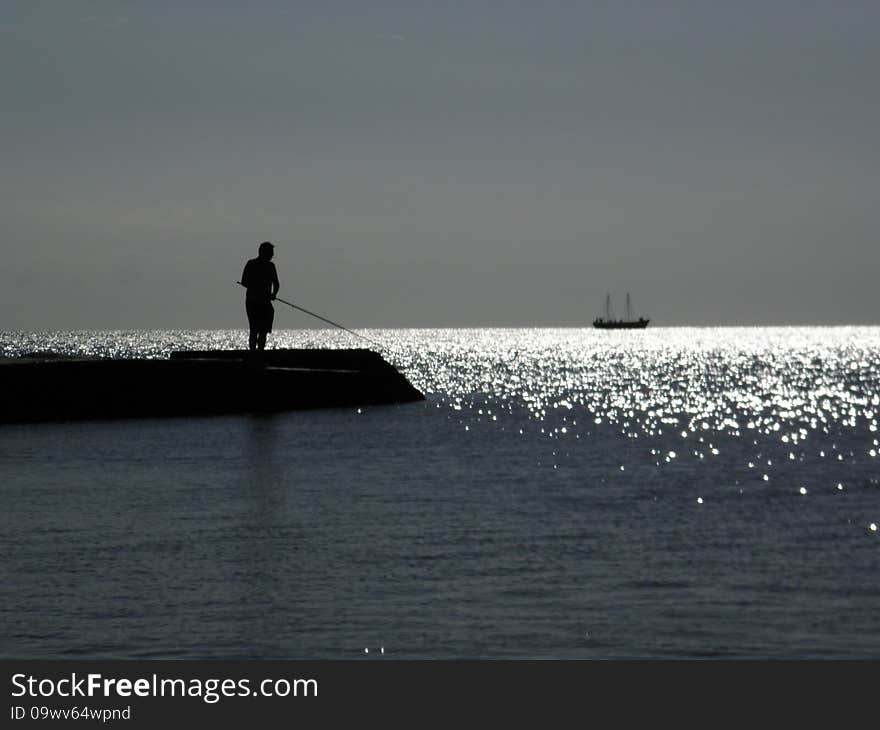 Figure of standing fisherman on a pier  on a background a sea and
swimming in the distance ship. Figure of standing fisherman on a pier  on a background a sea and
swimming in the distance ship