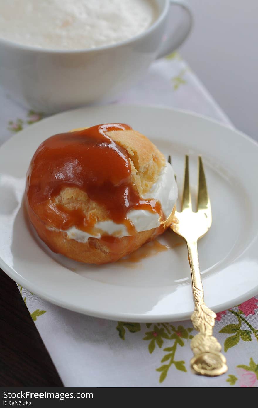 On a white plate one portion profiteroles with cream caramel sauce and a cup of cappuccino