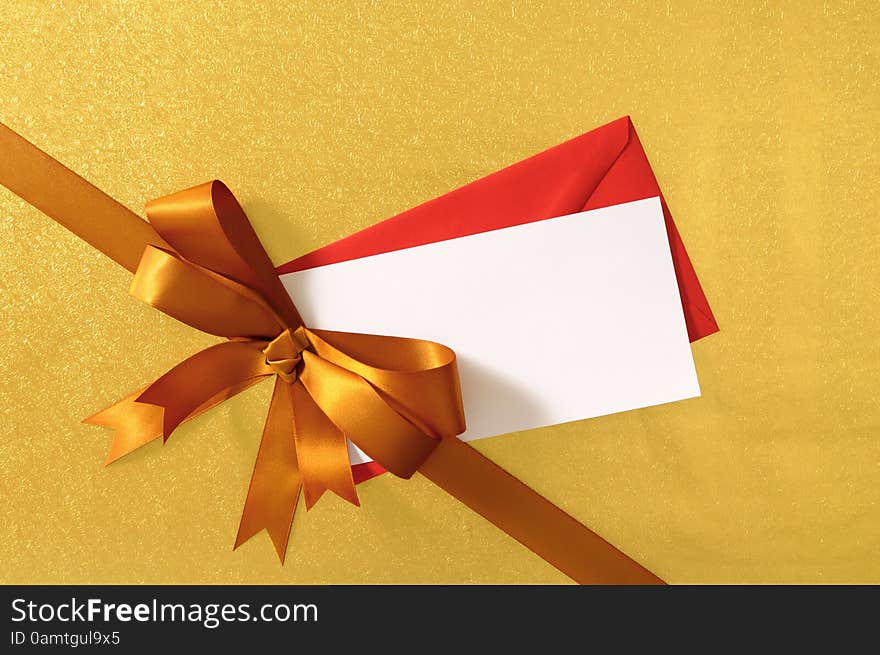 Christmas or birthday card with gift ribbon and bow in gold satin on shiny paper background with red envelope and blank message card. Christmas or birthday card with gift ribbon and bow in gold satin on shiny paper background with red envelope and blank message card