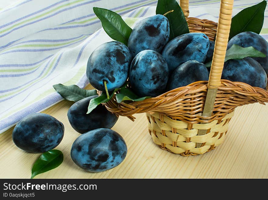 Ripe plums green leaves in small wicker basket on light wooden table with linen striped towel. Selective focus. Ripe plums green leaves in small wicker basket on light wooden table with linen striped towel. Selective focus