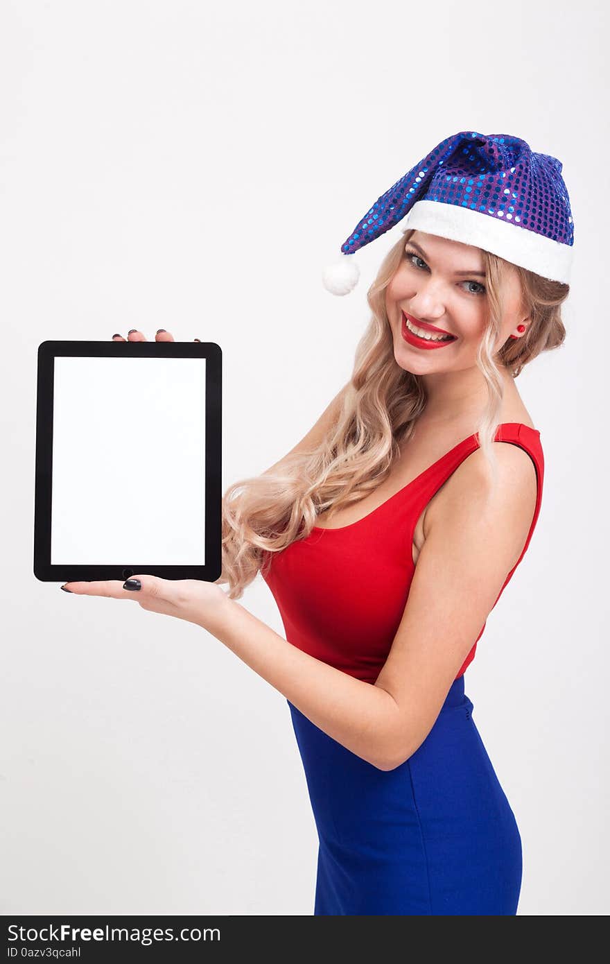 The beautiful blonde with big breasts standing on a white background and holds a a tablet in blue New Year's cap and smiling at the camera, selective focus on the tablet, picture with depth of field