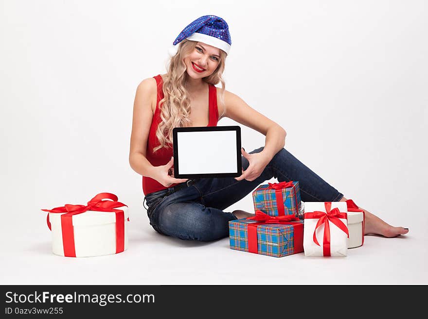 The beautiful blonde with big breasts is sitting on a white background and holds a a tablet in blue New Year's cap and smiling at the camera, selective focus on the tablet, picture with depth of field