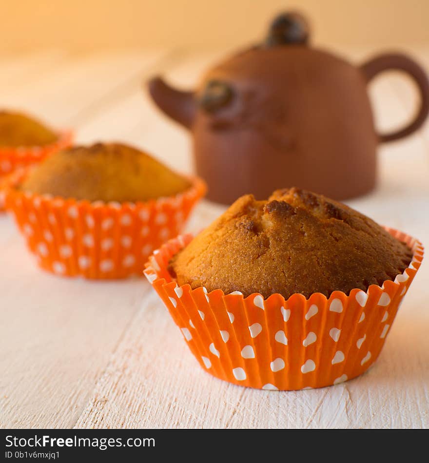 Muffins in orange paper cupcake holder with white polka dots and brown ceramic teapot on a white wooden background, selective focus, square. Muffins in orange paper cupcake holder with white polka dots and brown ceramic teapot on a white wooden background, selective focus, square