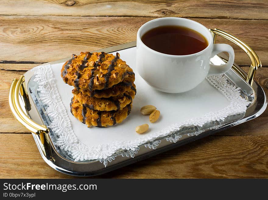 Chocolate icing cookies with peanuts and cup of tea on a metal serving tray with handles covered with a white lace napkin. Breakfast biscuits and tea.