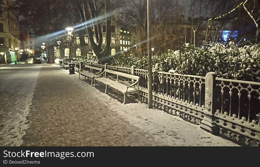 Snow covered fencing and bench on sidewalk and street illuminated in urban setting at night. Snow covered fencing and bench on sidewalk and street illuminated in urban setting at night.