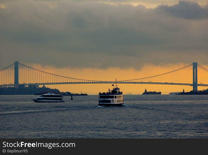 Boats on river by large suspension bridge against orange skies at sunset. Boats on river by large suspension bridge against orange skies at sunset.