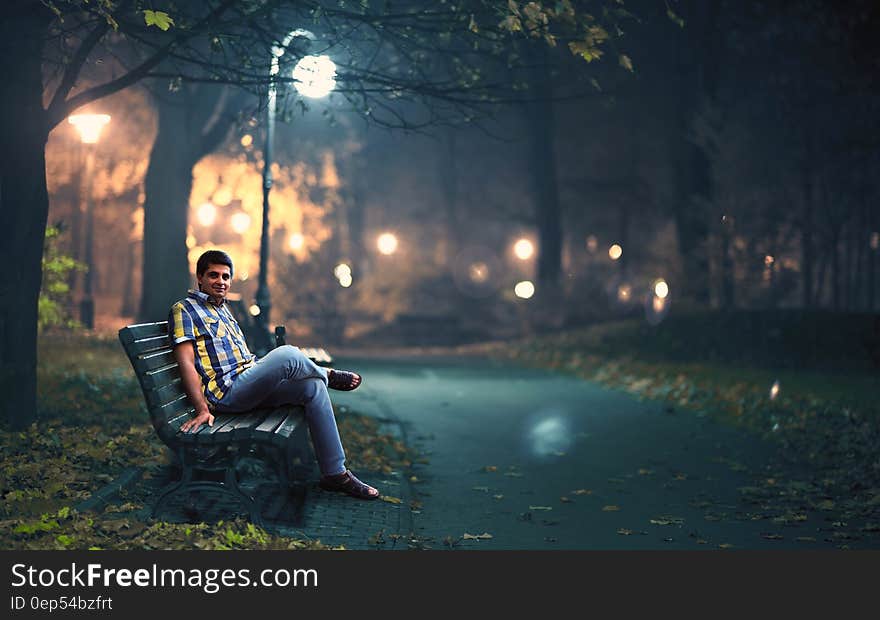 Man in Blue Denim Jeans Sitting Down by Wooden Bench Near Post Lamp Lighted