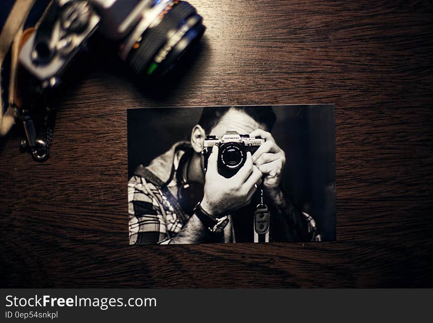 Still life of black and white snapshot of man photographing with Olympus camera next to camera on wooden table. Still life of black and white snapshot of man photographing with Olympus camera next to camera on wooden table.