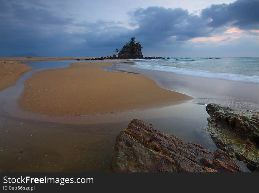 Sea Wave Crashing on Seashore With View of Rock Formation With Trees on Top of It Under Grey Clouds and White Sky
