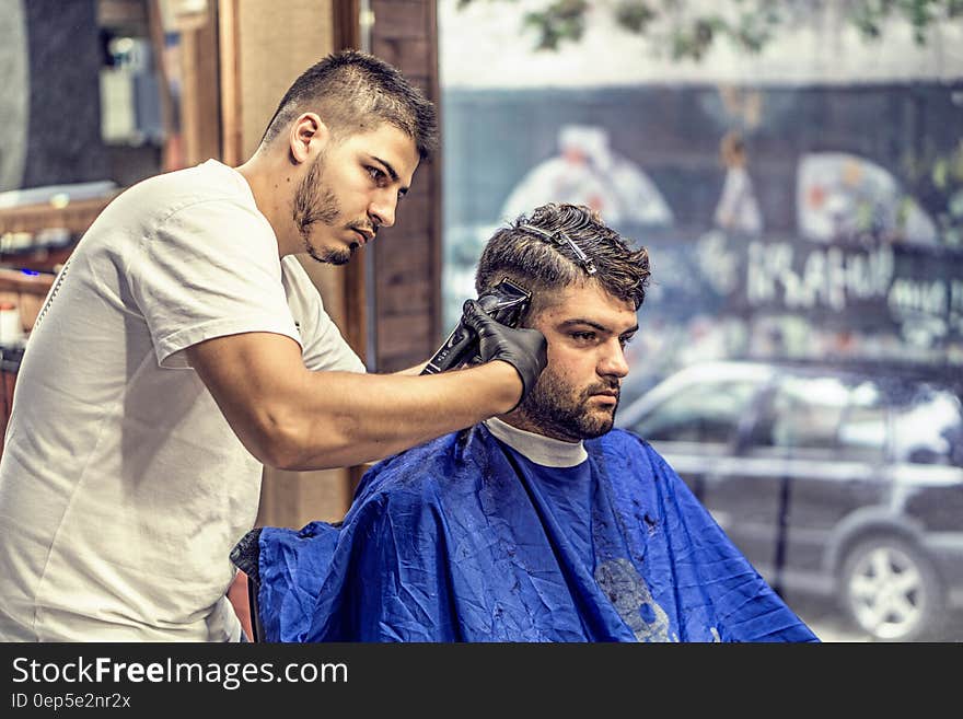 Barber in White Shirt Trimming Man&#x27;s Hair in Blue Textile While Sitting Nearby Glass Window