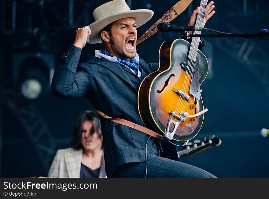 Man in Black Formal Suit Holding Electric Guitar While Raising His Right Hand and Opening His Mouth in Tilt Shift Lens Photography