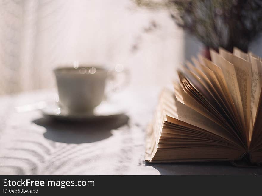 Tilt Lens Photography of Open Book With Ceramic Cup in Saucer