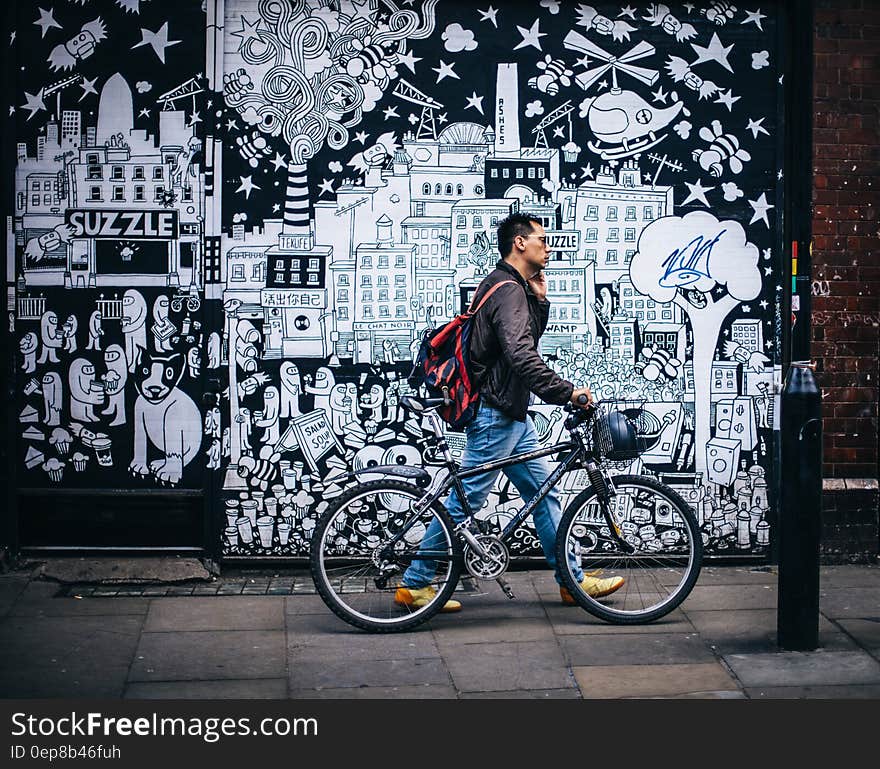 Man in Black Jacket Holding a Black Hardtail Bike Near Black and White Art Wall