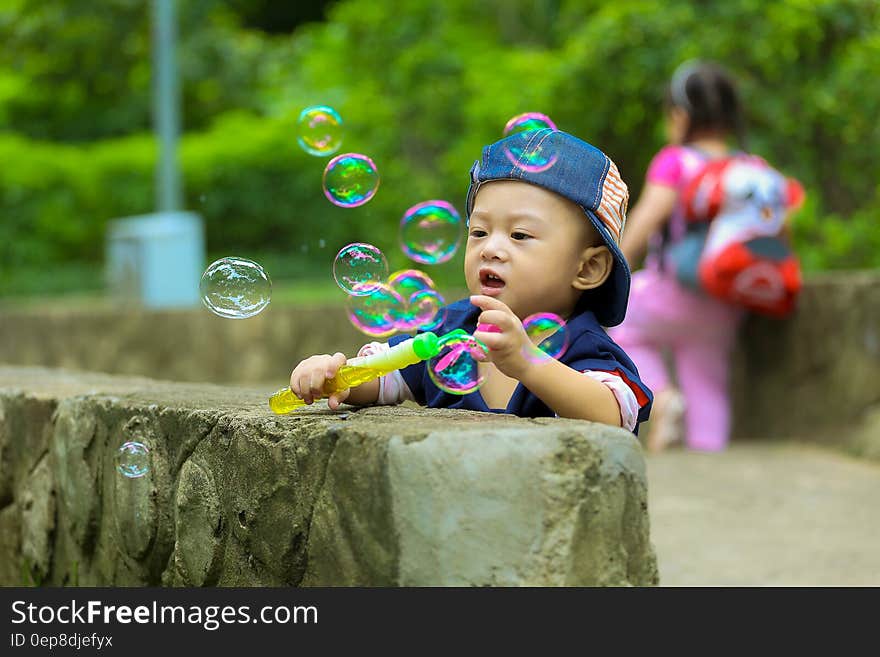 Boy in Blue Fitted Cap Playing Bubbles and Leaning on Grey Concrete Wall at Daytime