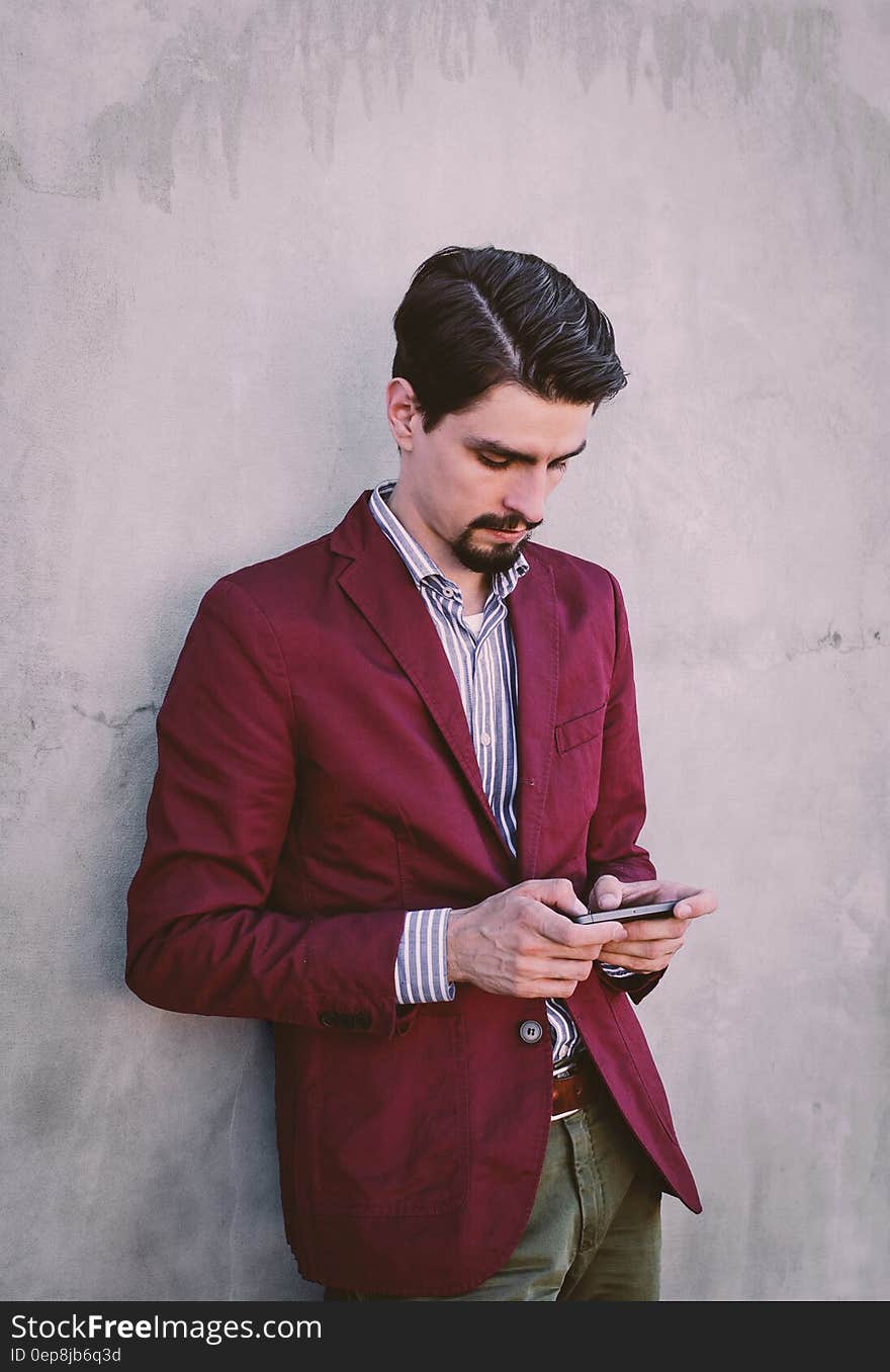 Man Wearing Maroon Blazer Leaning on Gray Concrete Wall While Using His Smartphone