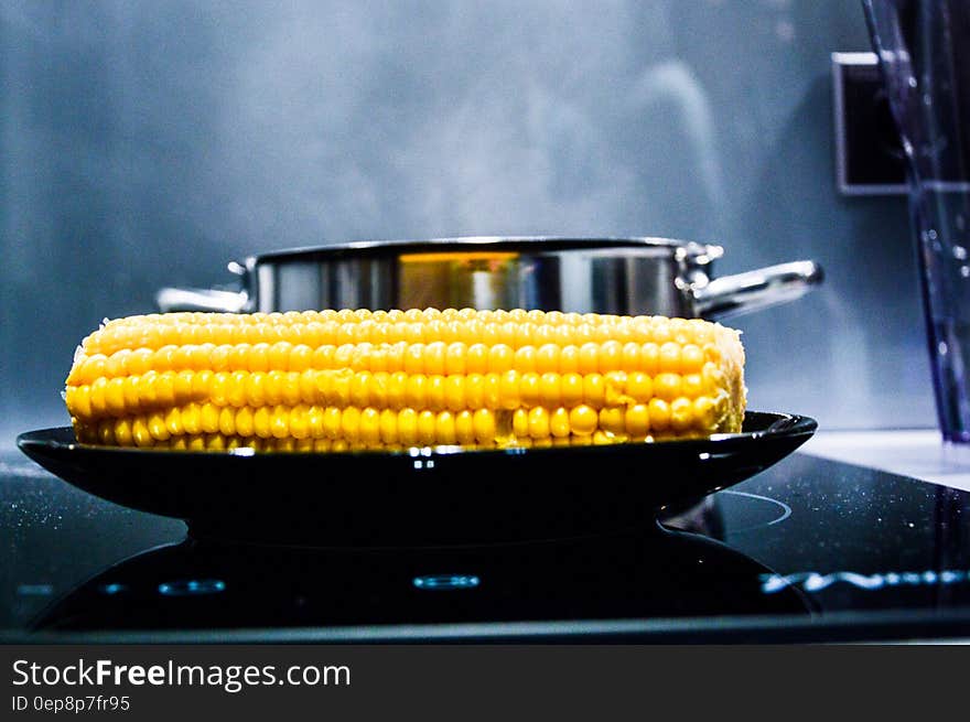 Plate of corn on the cob next to steaming metal pot in kitchen.