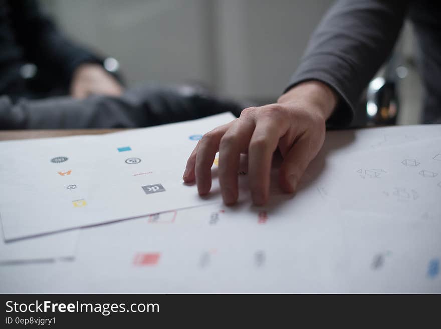 Designer with a hand on sheets of free sample icons from which he must choose (select), office background. Designer with a hand on sheets of free sample icons from which he must choose (select), office background.