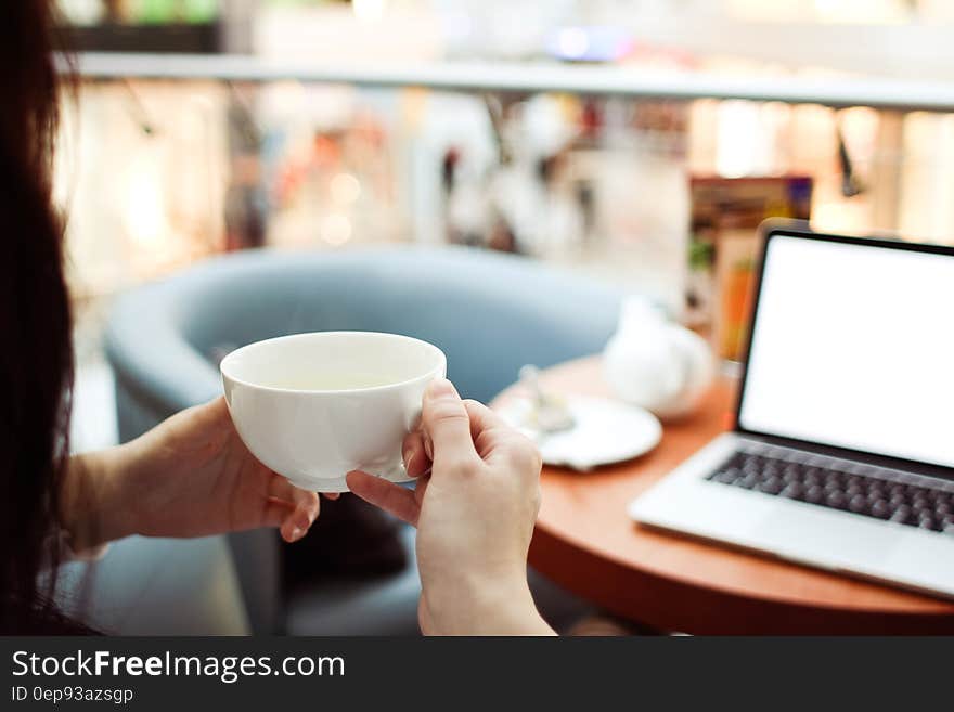 Person Holding White Ceramic Teacup in Front of a Macbook Pro