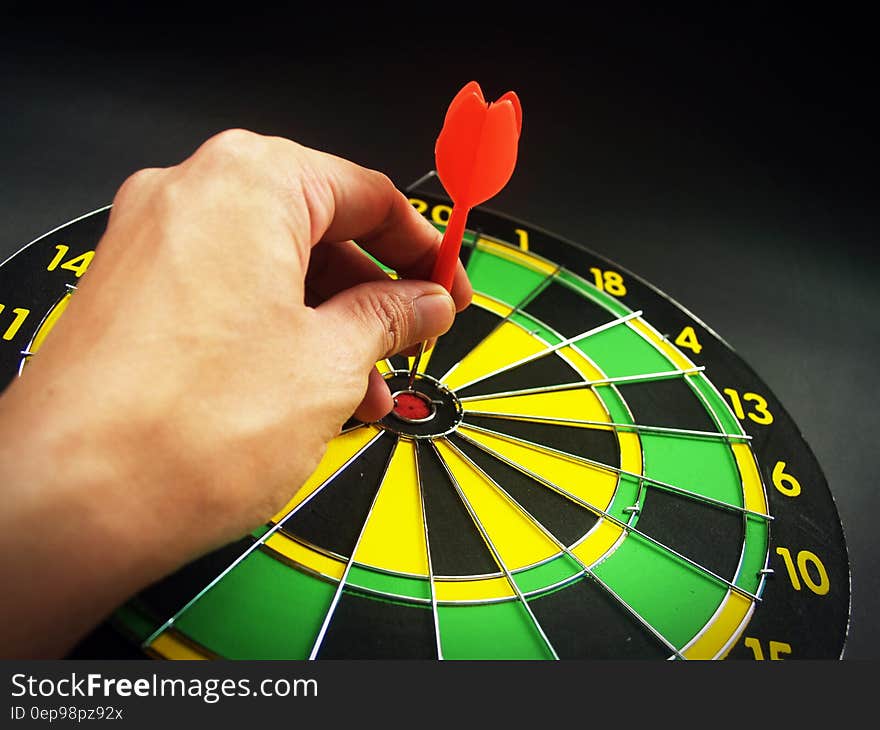 Person Holding Red Dart on Green Yellow and Black Dart Board