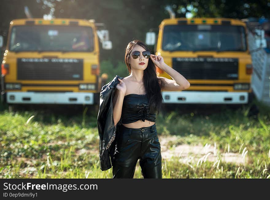 Glamor portrait of dark haired woman posing in black pants and top holding jacket wearing sunglasses outdoor with yellow trucks in field. Glamor portrait of dark haired woman posing in black pants and top holding jacket wearing sunglasses outdoor with yellow trucks in field.