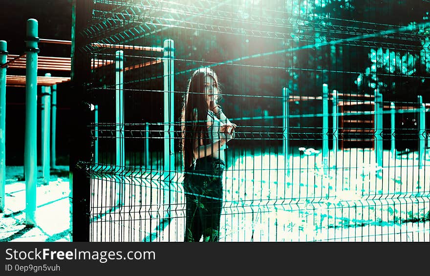 Fashionable young girl stood behind a fence in outdoors playground with bright turquoise lighting highlights.