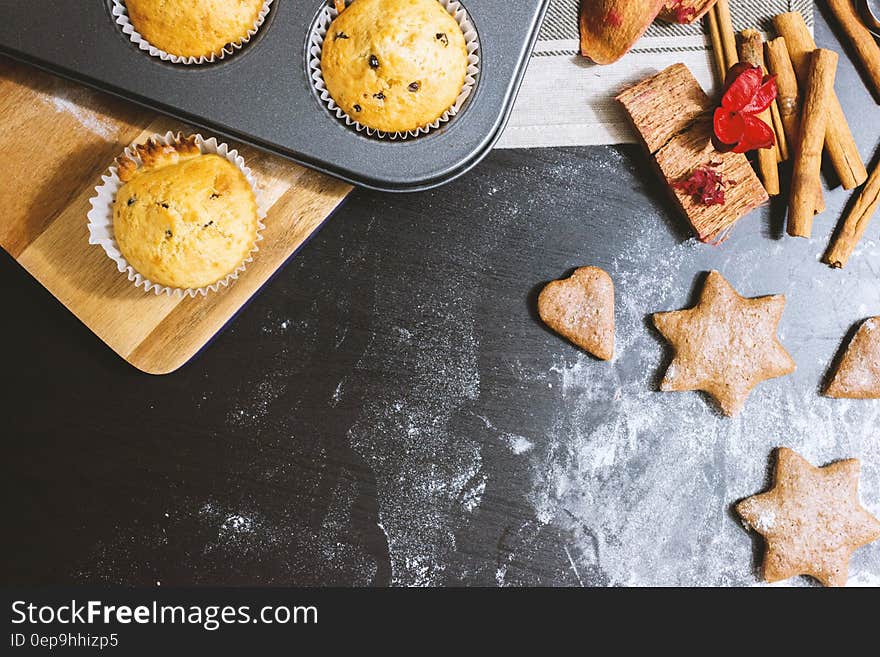 Surface with flour and star shaped biscuits and next to that cup cakes with currents. Other items include cinnamon sticks and a red decorative butterfly are placed alongside. Surface with flour and star shaped biscuits and next to that cup cakes with currents. Other items include cinnamon sticks and a red decorative butterfly are placed alongside.