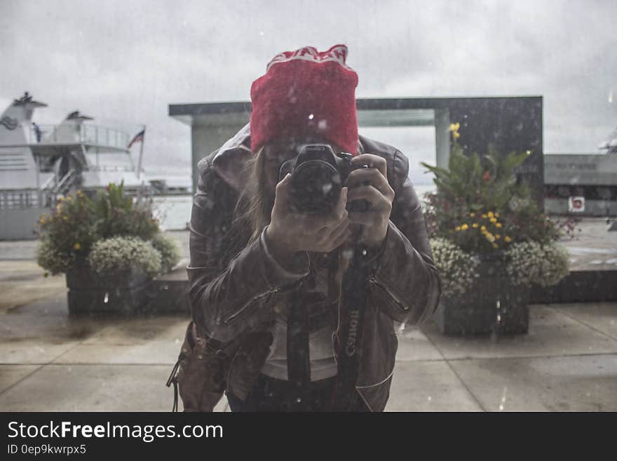 Woman in Brown Leather Jacket and Red Knit Cap Using Black Dslr Camera