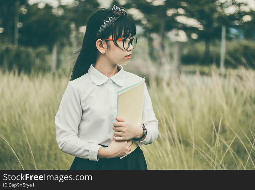 Young attractive Asian schoolgirl with white blouse, dark skirt and spectacles holding school folder while standing outdoors in a hay field, background of dark green trees. Young attractive Asian schoolgirl with white blouse, dark skirt and spectacles holding school folder while standing outdoors in a hay field, background of dark green trees.