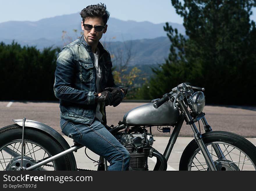 Young man in leather jacket and blue jeans sitting astride a stationary vintage style motorbike with spoke wheels, landscape of dense forest and distant blue mountains. Young man in leather jacket and blue jeans sitting astride a stationary vintage style motorbike with spoke wheels, landscape of dense forest and distant blue mountains.