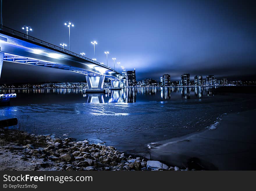 Modern bridge illuminated at night from waterfront with urban skyline reflecting in waters. Modern bridge illuminated at night from waterfront with urban skyline reflecting in waters.