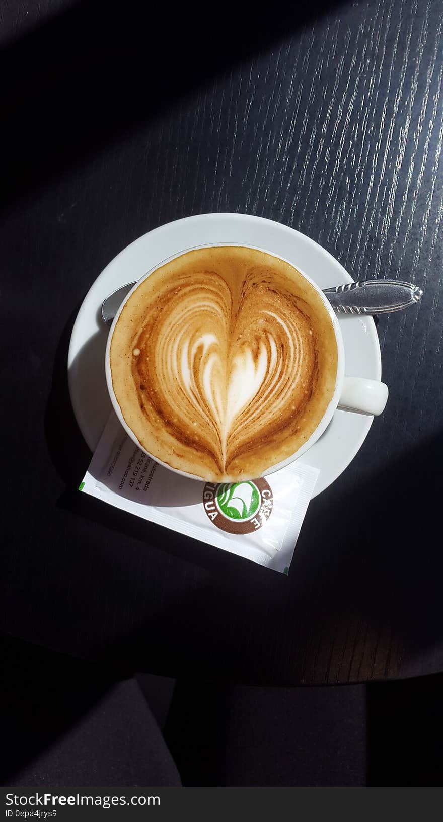 Overhead view of white china cup and saucer with cappuccino foam with design and spoon on wooden tabletop. Overhead view of white china cup and saucer with cappuccino foam with design and spoon on wooden tabletop.