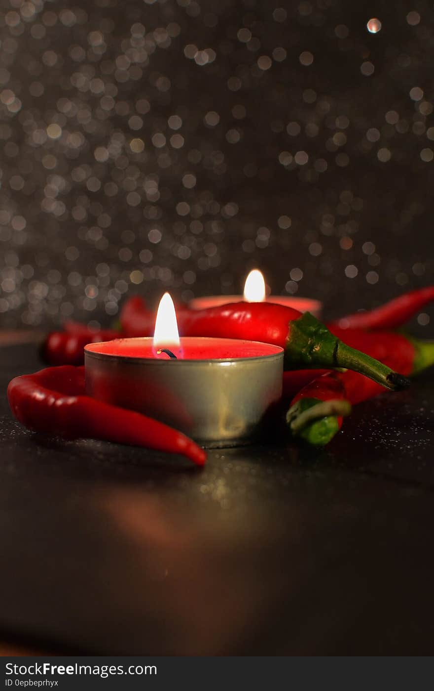 Two burning candles surrounded by red chili peppers on a table. Two burning candles surrounded by red chili peppers on a table.