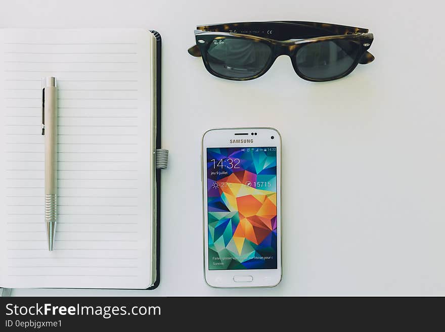 White Samsung Smartphone Beside Sunglasses,pen and White Notebook