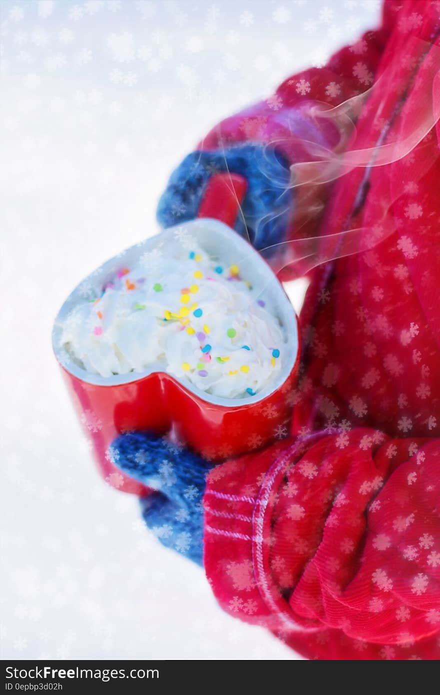 Person in red jacket and blue gloves holding red heart shaped mug of cocoa with whipped cream and sprinkles with snowflakes. Person in red jacket and blue gloves holding red heart shaped mug of cocoa with whipped cream and sprinkles with snowflakes.