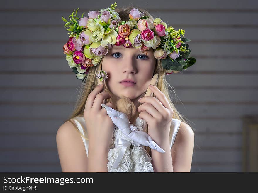 Portrait of young pretty girl wearing floral head-dress composed of many different flowers, white dress with bow and looking pensive, gray slats for a background. Portrait of young pretty girl wearing floral head-dress composed of many different flowers, white dress with bow and looking pensive, gray slats for a background.