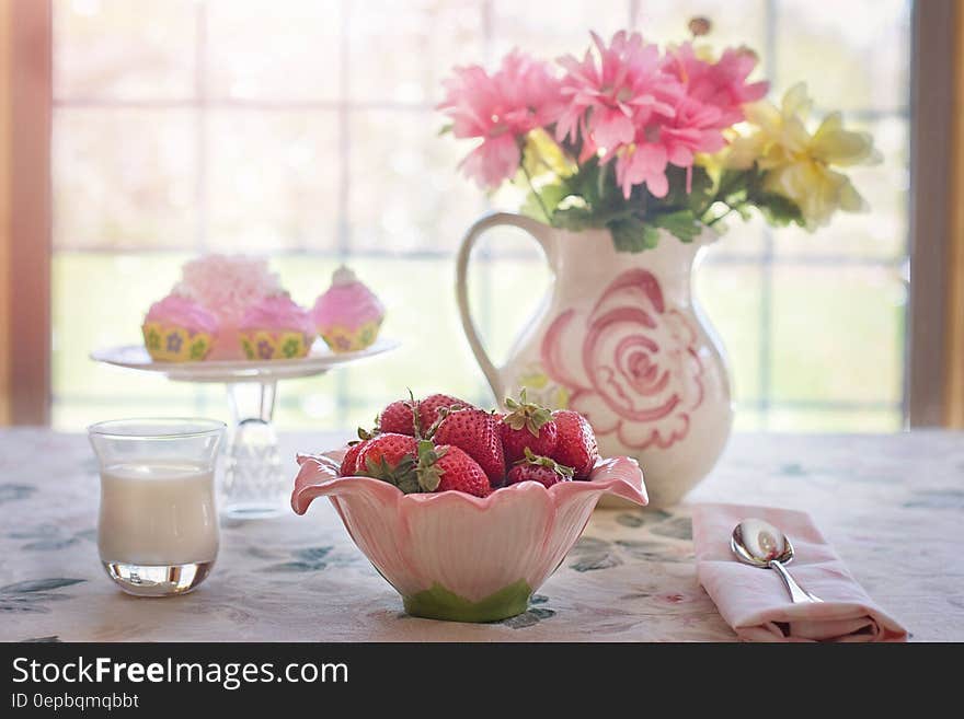 Still life on tabletop with strawberries in pink bowl, glass of milk or cream, plate of cupcakes and vase of flowers against sunny window. Still life on tabletop with strawberries in pink bowl, glass of milk or cream, plate of cupcakes and vase of flowers against sunny window.