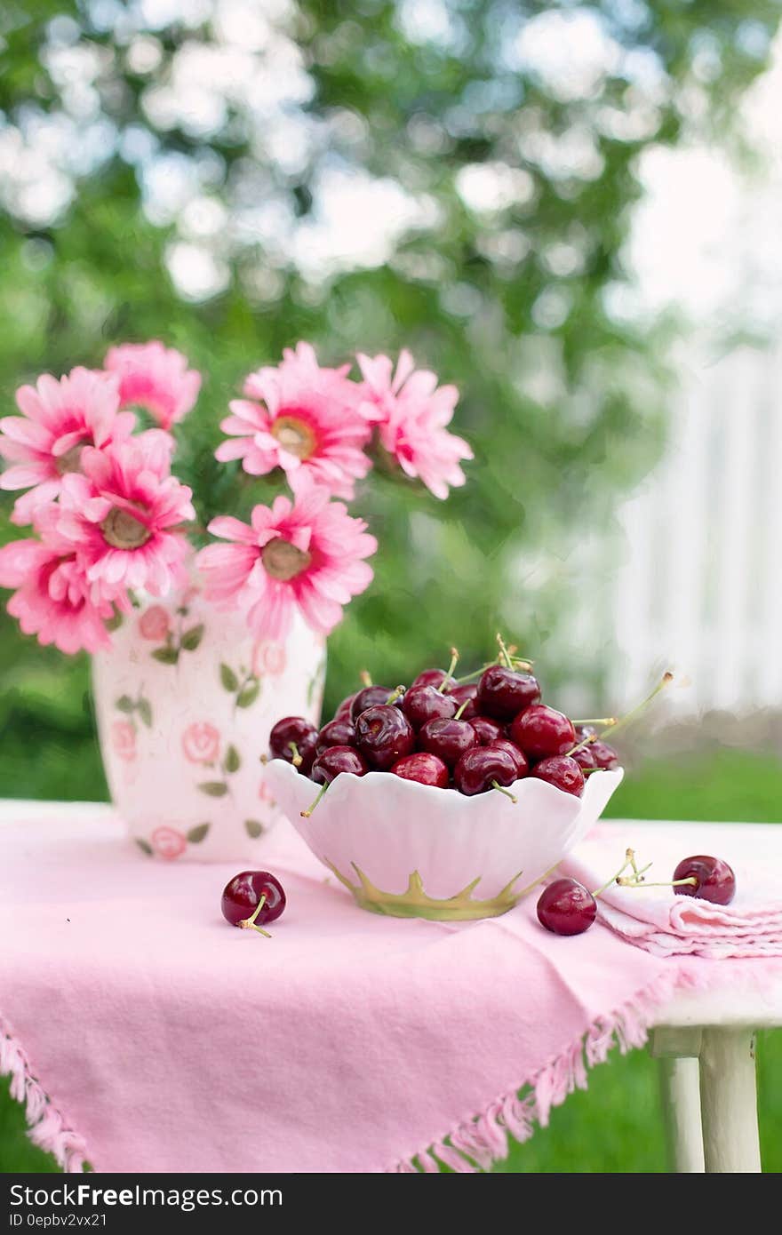 Pink Petaled Flower Beside White and Green Bowl Full of Cherry