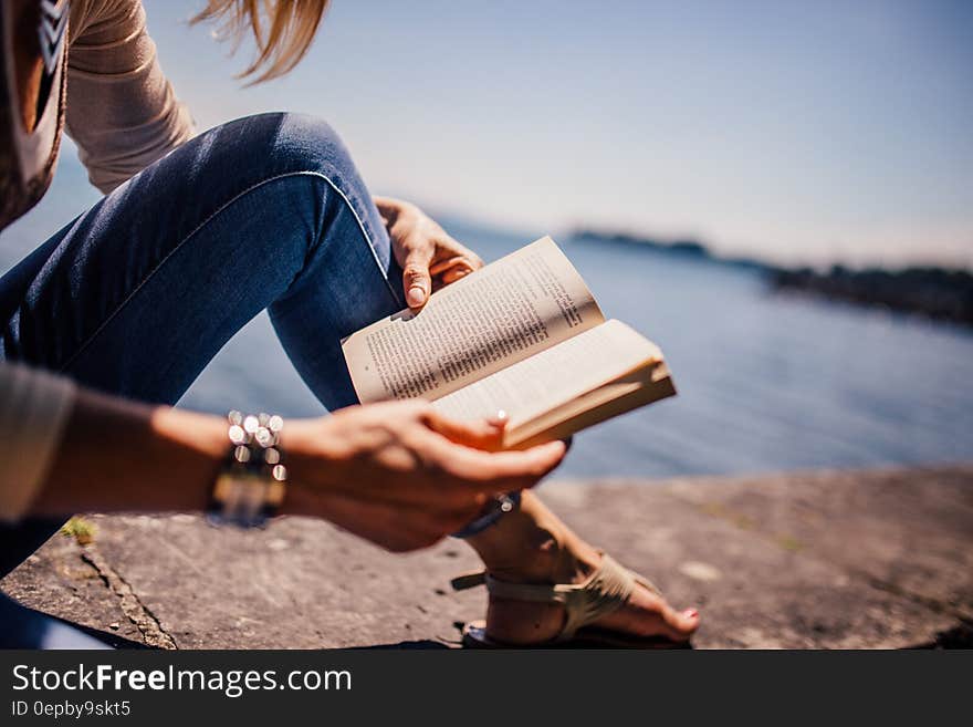 Woman Wearing Blue Denim Jeans Holding Book Sitting on Gray Concrete at Daytime