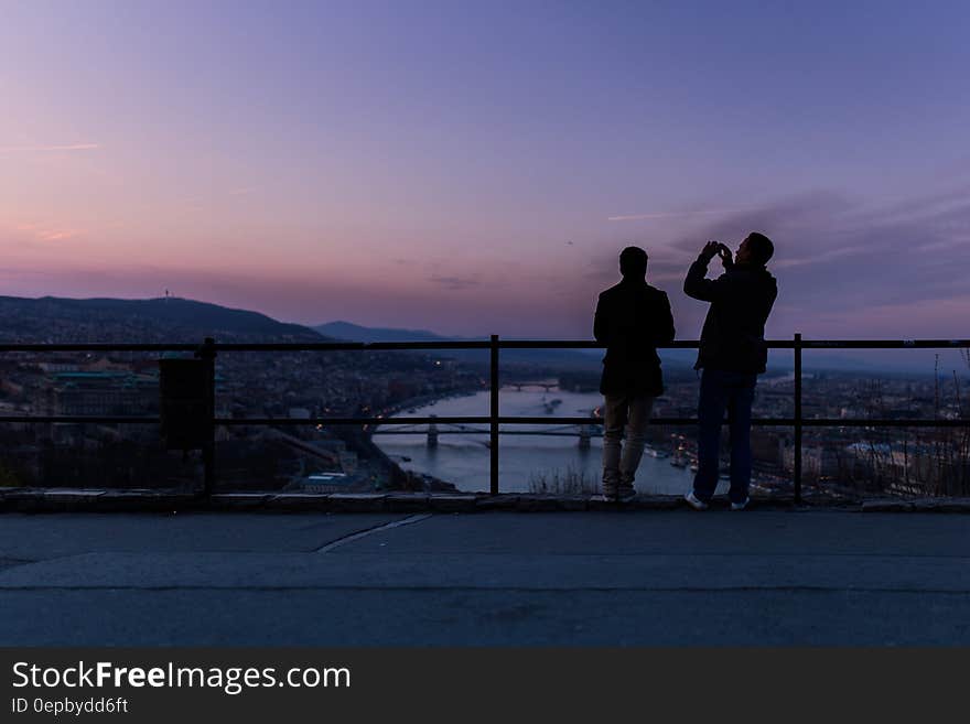 A couple of men standing by a fence looking down into a city. A couple of men standing by a fence looking down into a city.